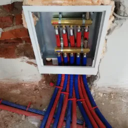 Rozdzielacz instalacji ogrzewania podłogowego w skrzynce, z widocznymi rurami doprowadzającymi ciepłą i zimną wodę, zamontowany w surowym otworze w ścianie.