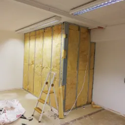 Montaż izolacji z wełny mineralnej w metalowej konstrukcji szkieletowej ściany działowej, widoczna drabina malarska i resztki materiałów budowlanych w workach na podłodze.