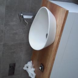 Krzysztof Mielnik Remonty i Wykończenia - Nowoczesna, owalna umywalka nablatowa zamontowana na drewnianym blacie, z minimalistyczną baterią i szarą płytką na ścianie. Widoczny fragment szafki pod umywalką w kolorze białym i pozostałości po...