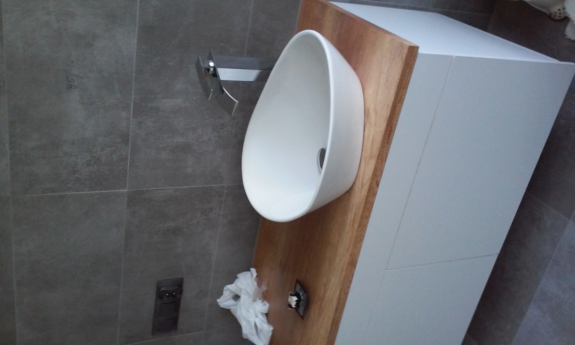 Nowoczesna, owalna umywalka nablatowa zamontowana na drewnianym blacie, z minimalistyczną baterią i szarą płytką na ścianie. Widoczny fragment szafki pod umywalką w kolorze białym i pozostałości po...