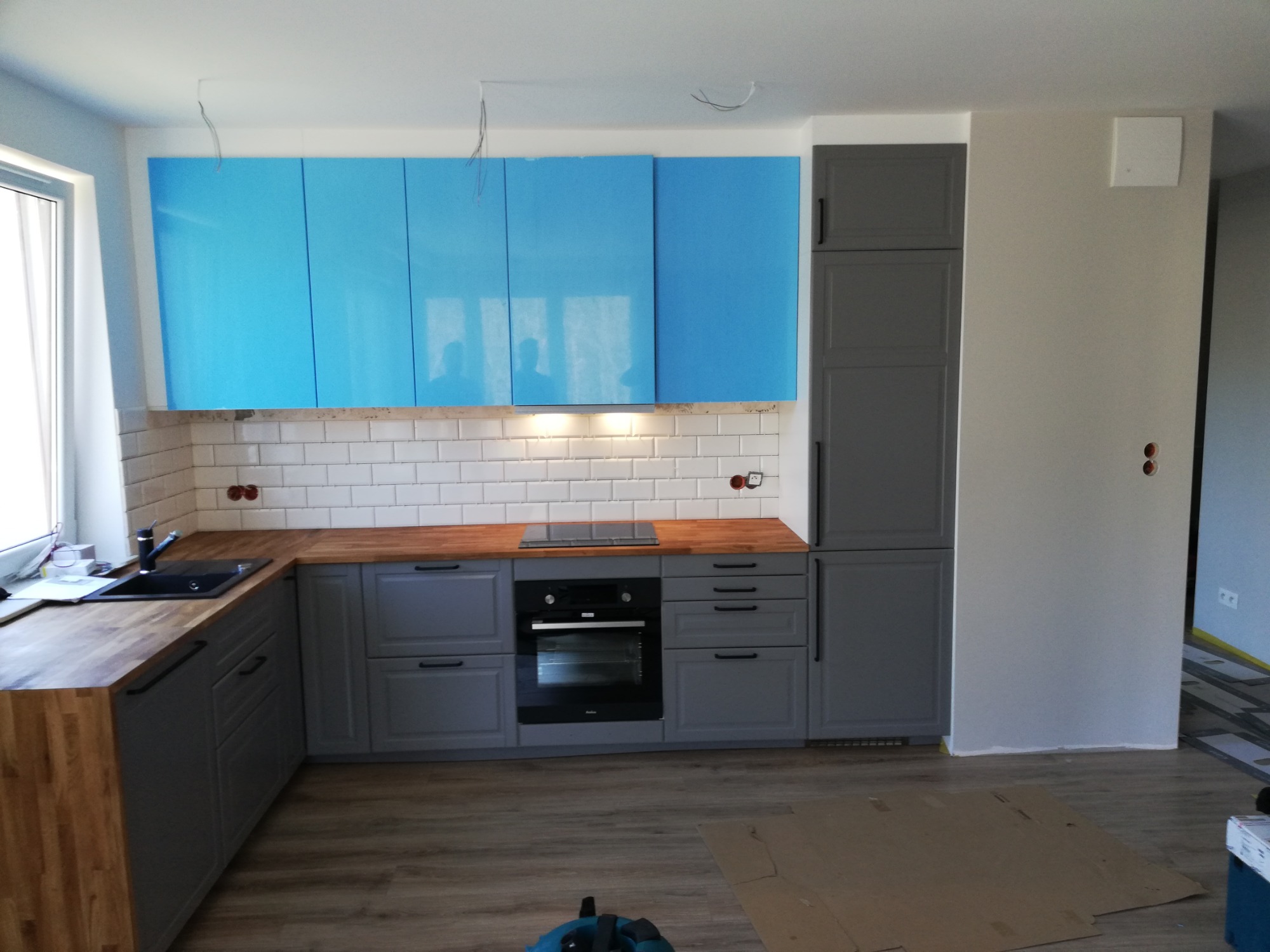 Szara zabudowa kuchenna z drewnianym blatem i turkusowymi szafkami wiszącymi, białe płytki na ścianie, widoczne niedokończone podłączenia elektryczne.