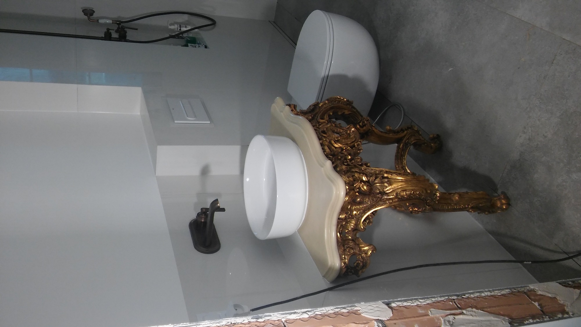 Nowoczesna biała umywalka z ceramiczną misą i złotą, bogato zdobioną podstawą w łazience z białymi płytkami i szarą podłogą, obok toaleta.