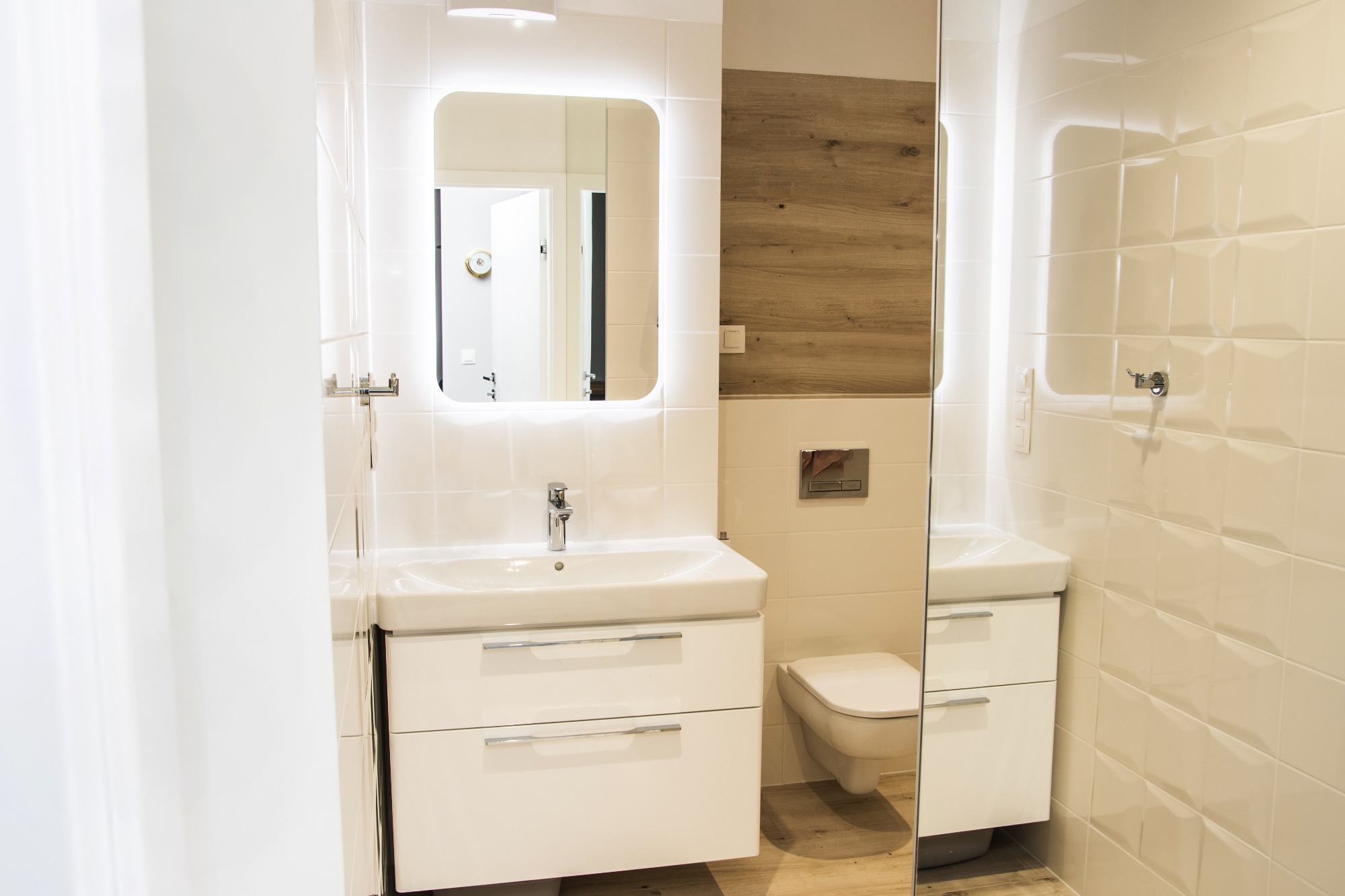 Nowoczesna, jasna łazienka z białą armaturą i płytkami, podświetlanym lustrem oraz akcentem drewna na ścianie.