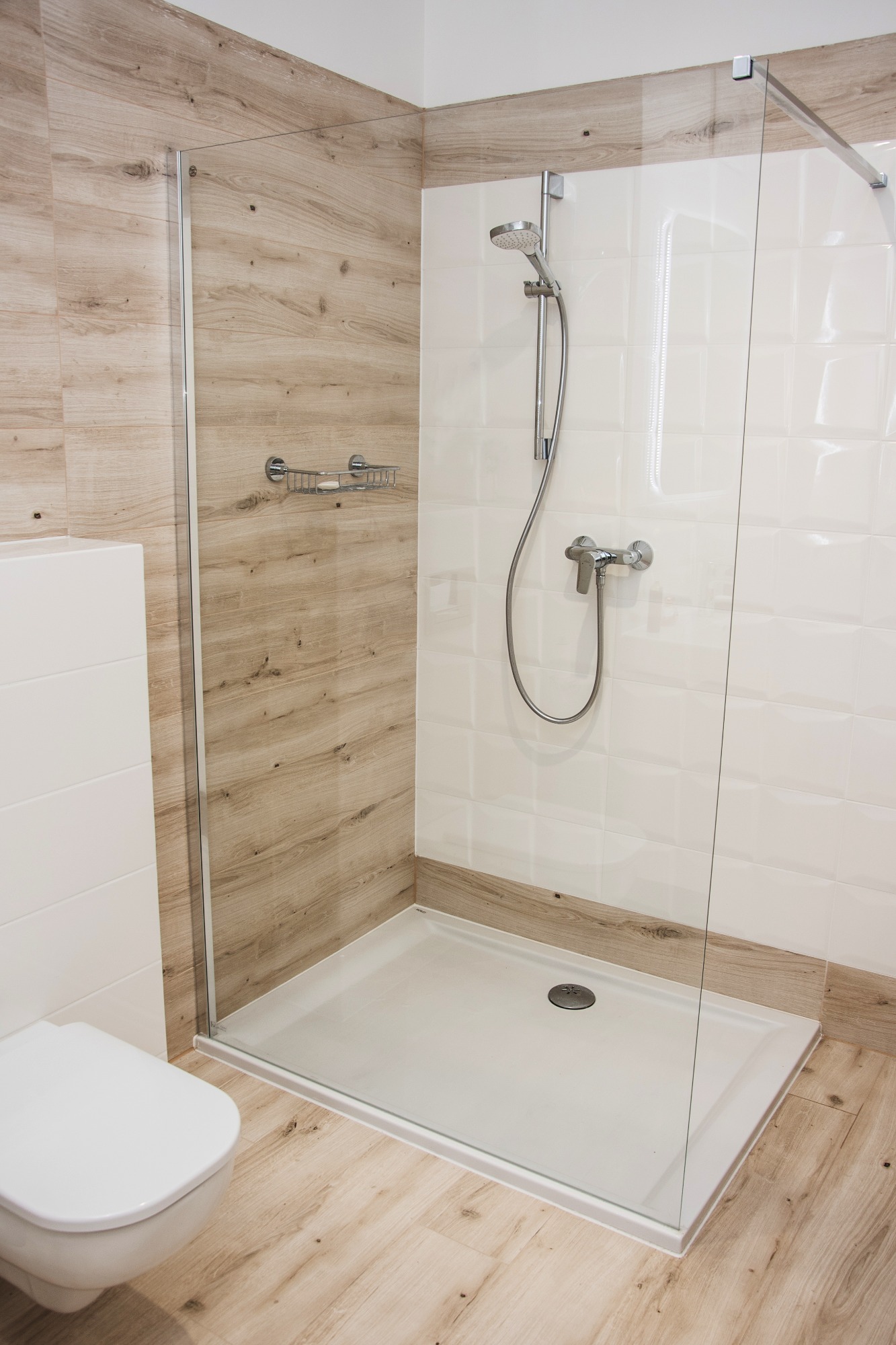 Nowoczesna kabina prysznicowa z drewnopodobnymi płytkami na ścianach, szklaną ścianką i białą armaturą, widok z boku na toaletę.