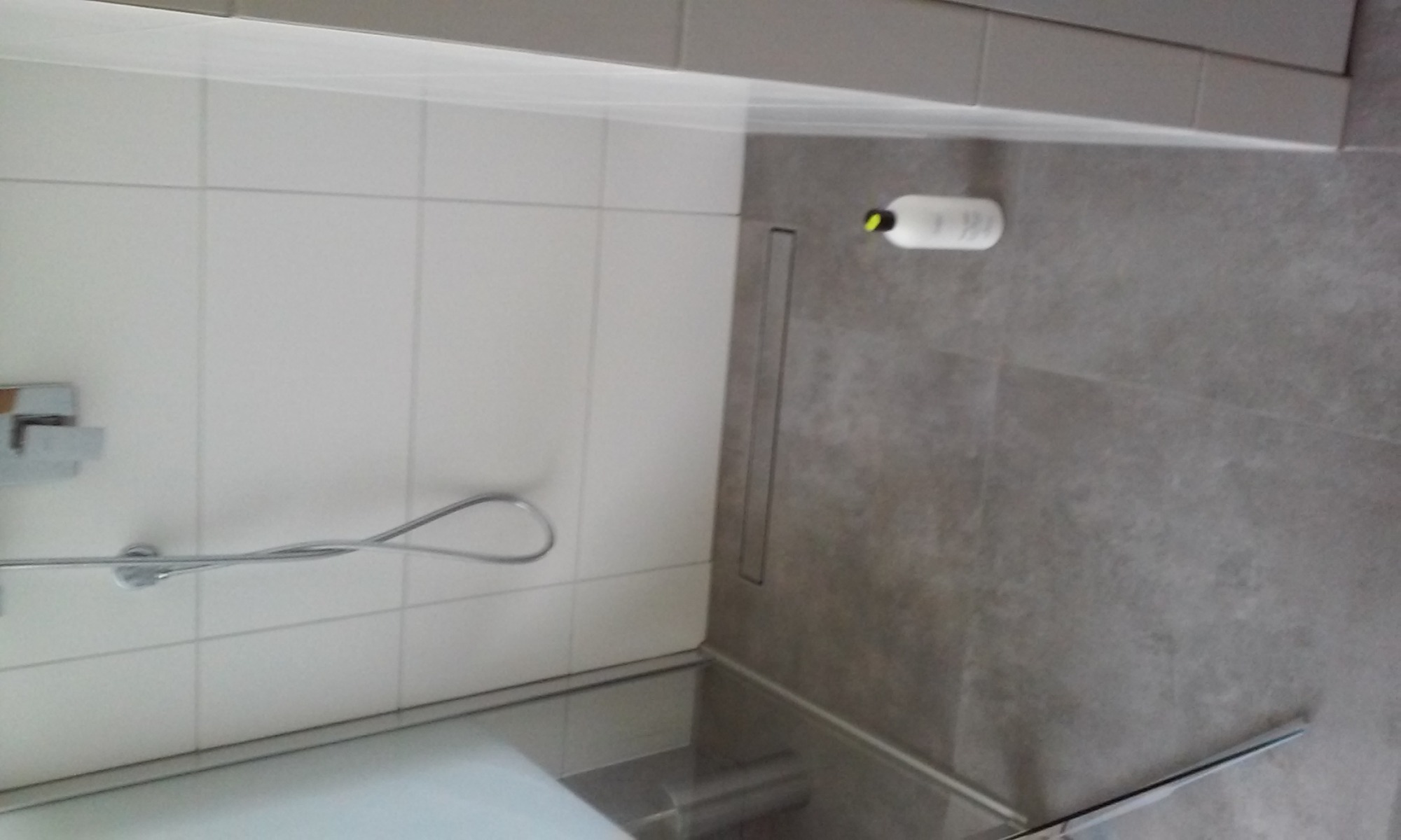 Wykończona łazienka z białymi płytkami na ścianach, szarymi płytkami na podłodze, odpływem liniowym i butelką szamponu leżącą na podłodze.