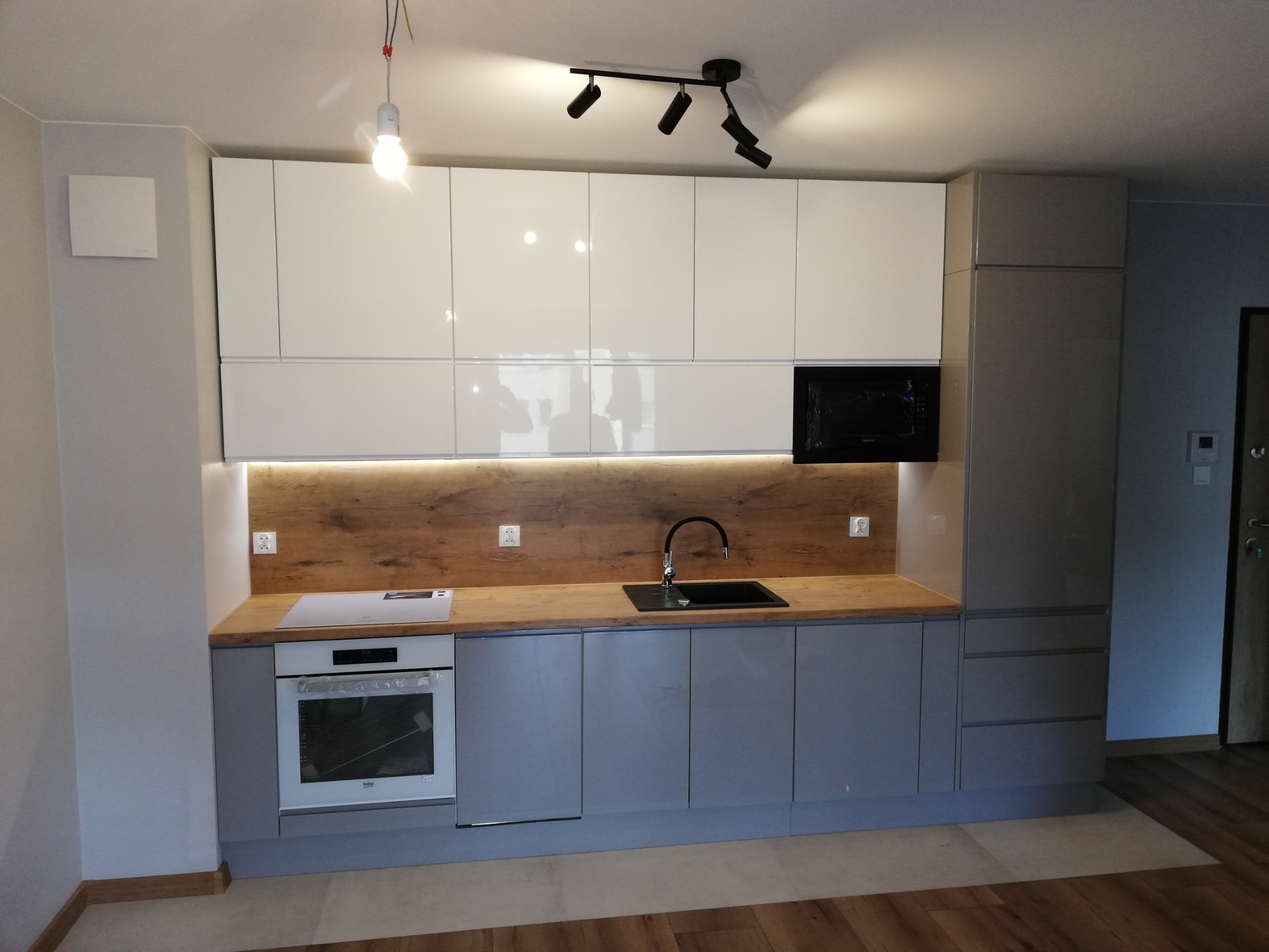 Nowoczesna kuchnia z białymi górnymi szafkami na wysoki połysk, szarymi dolnymi szafkami i drewnianym blatem z oświetleniem LED podszafkowym. Widoczny czarny zlew, płyta indukcyjna i piekarnik.