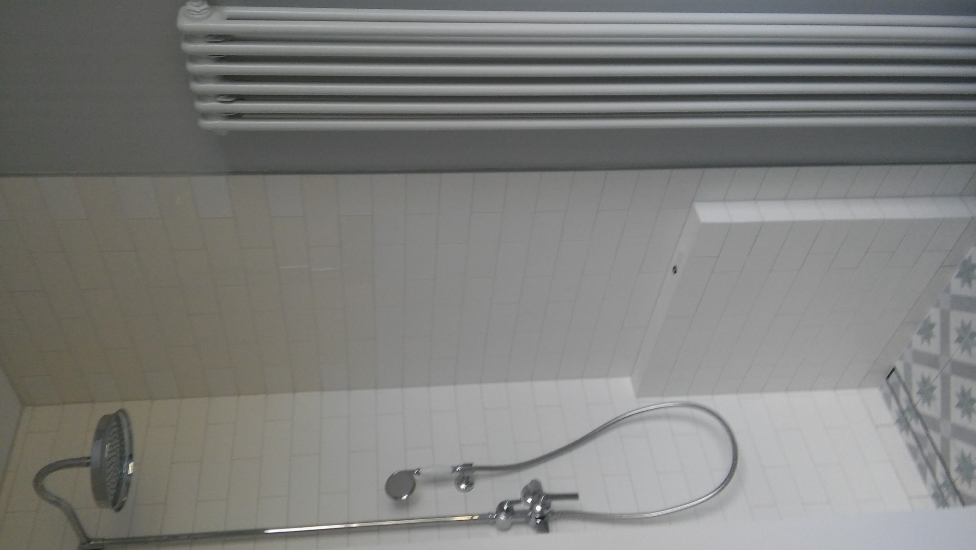 Wnętrze łazienki z białymi, prostokątnymi płytkami ułożonymi w cegiełkę, widoczna bateria prysznicowa z deszczownicą, grzejnik drabinkowy pod sufitem i fragment dekoracyjnej ściany z szarymi wzorami.