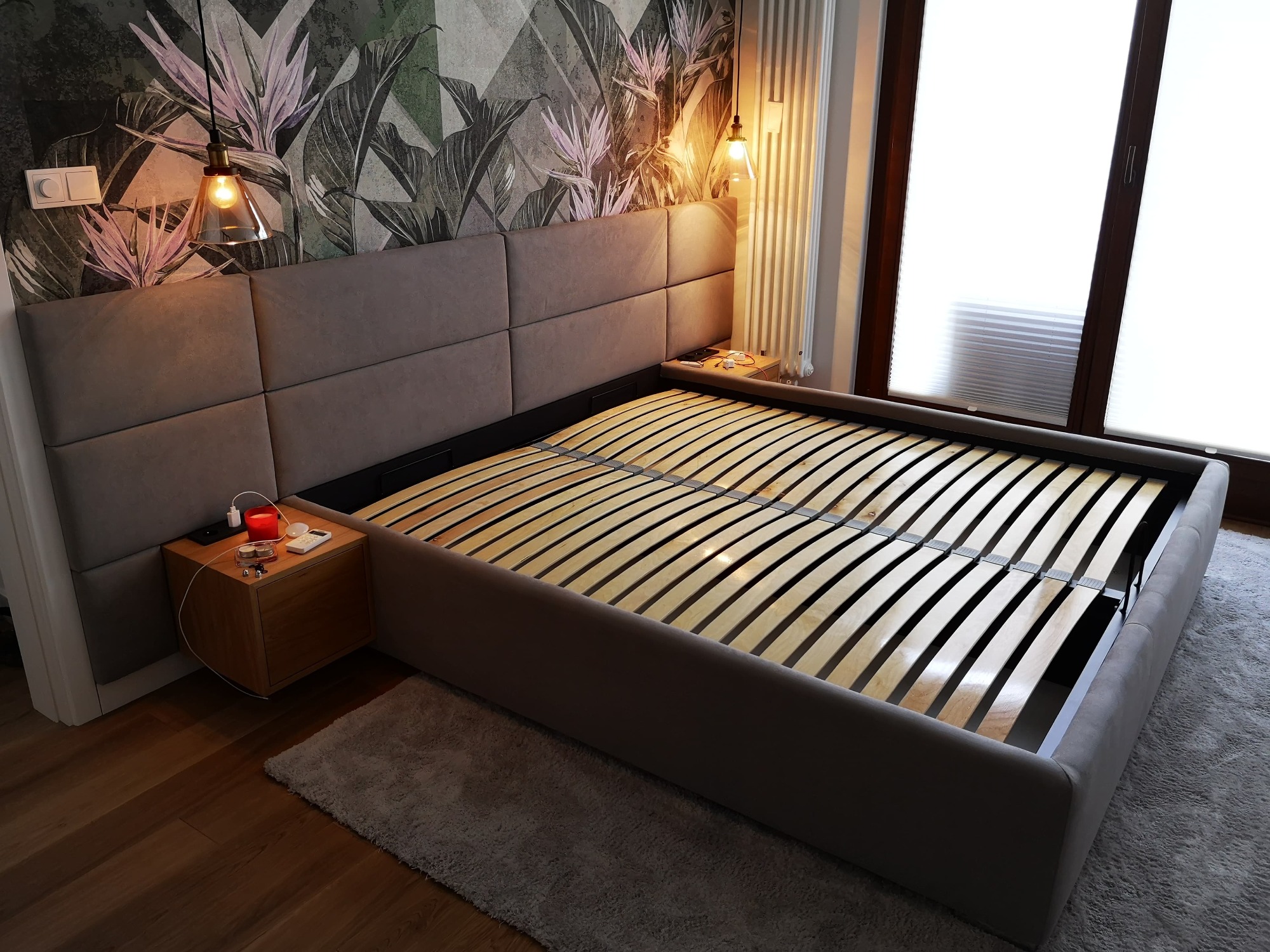 Szare łóżko z tapicerowanym wezgłowiem w kwadraty i drewnianym stelażem, obok szafka nocna z zapaloną świeczką i ładowarkami, na tle tapeta w liście i okno z żaluzjami.