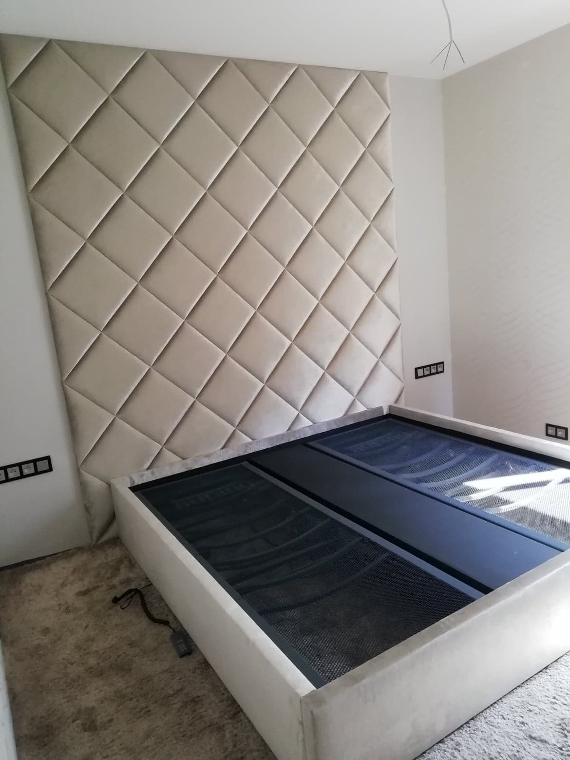 Beżowe tapicerowane wezgłowie łóżka z geometrycznym wzorem rombów, widoczny stelaż z czarną siatką, w tle biała ściana z gniazdkami elektrycznymi.