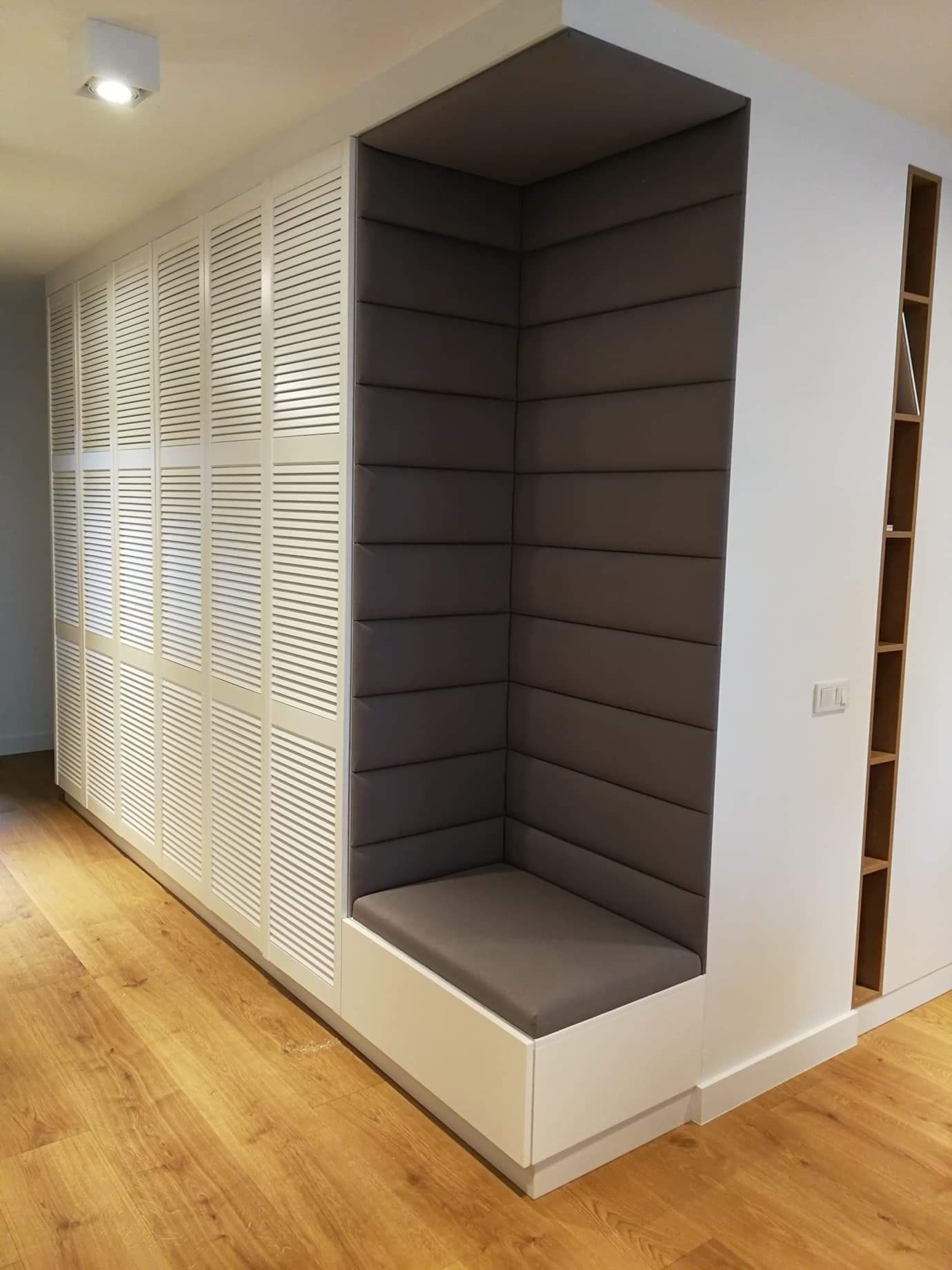 Biała szafa z lamelowymi drzwiami i wnęką z tapicerowanym siedziskiem w kolorze szarym, na drewnianej podłodze.