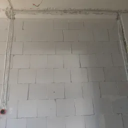 Ściana z betonu komórkowego z widocznymi, nowo położonymi przewodami elektrycznymi w białych rurkach, biegnącymi wzdłuż ściany i pod sufitem. Widoczne puszki elektryczne w kolorze pomarańczowym.