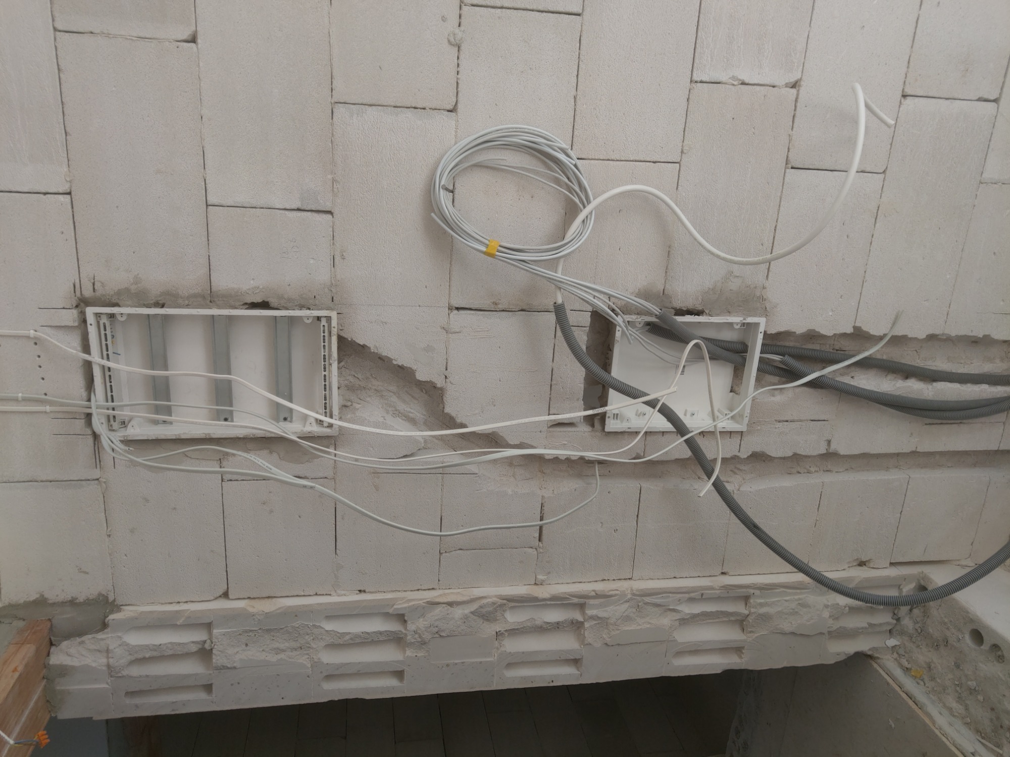 Dwa puste otwory pod gniazdka elektryczne w ścianie z betonu komórkowego, z widocznymi przewodami elektrycznymi, w tym zwój kabla zabezpieczony żółtą taśmą.
