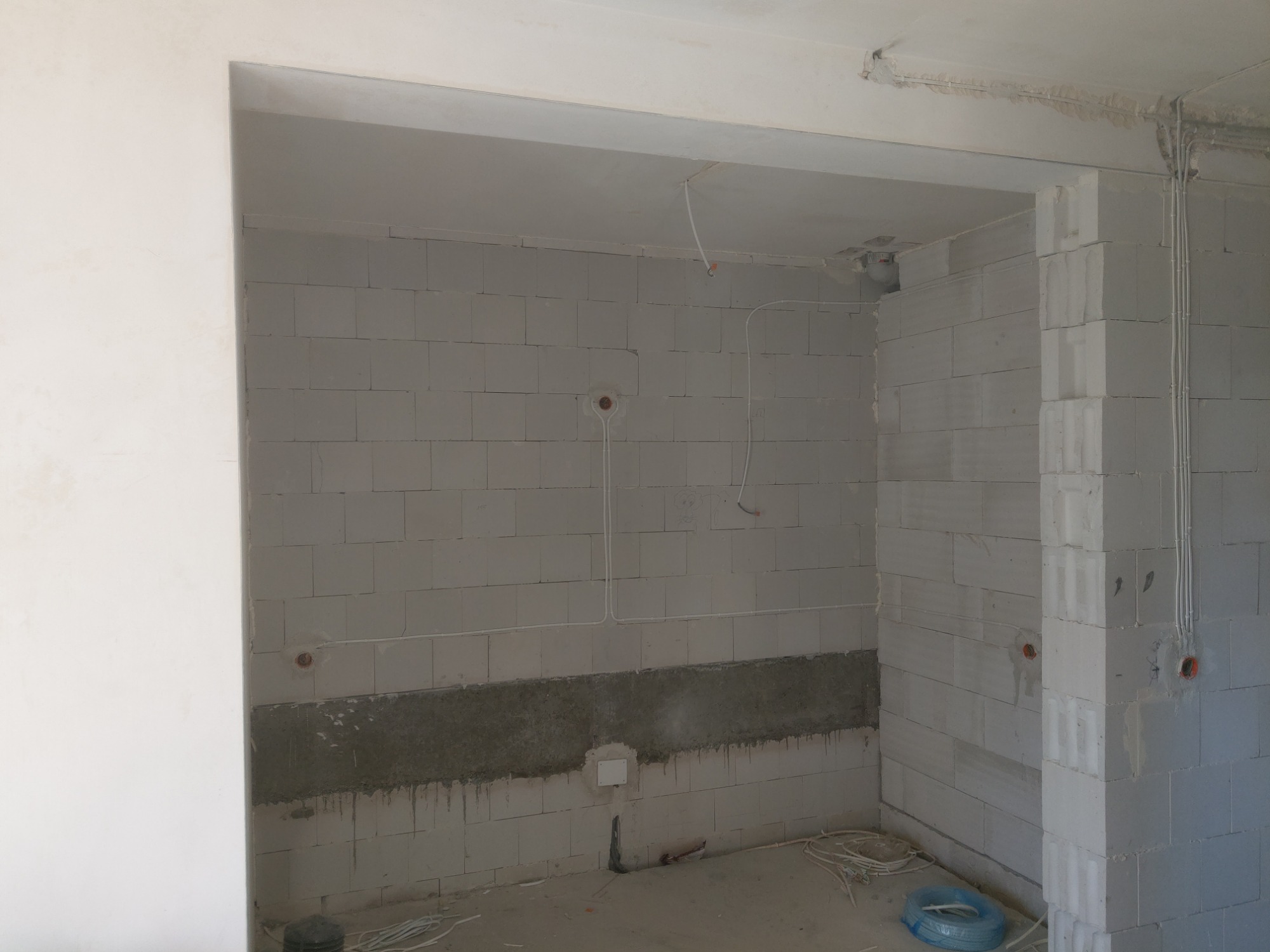 Surowy stan wnętrza z białymi ścianami z betonu komórkowego, widoczne przewody elektryczne w ścianach i suficie, częściowo wykończona ściana z szarym pasem. Na podłodze leżą zwinięte kable.
