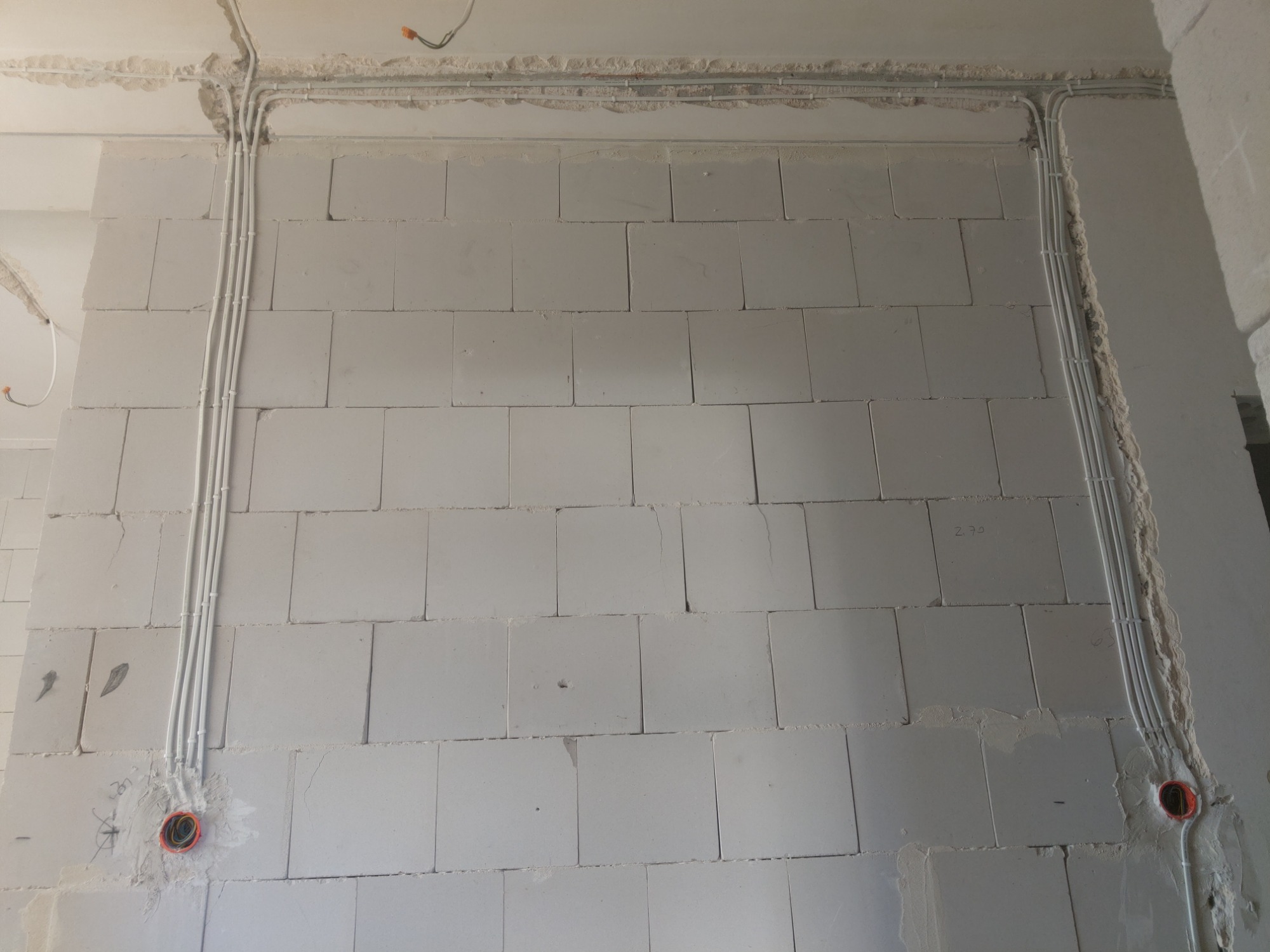 Ściana z betonu komórkowego z widocznymi, nowo położonymi przewodami elektrycznymi w białych rurkach, biegnącymi wzdłuż ściany i pod sufitem. Widoczne puszki elektryczne w kolorze pomarańczowym.