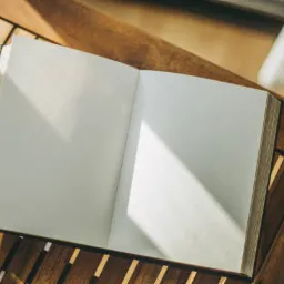 Otwarta księga z pustymi stronami leży na drewnianym stoliku w słoneczny dzień w Katowicach. Promień światła pada na prawą stronę rozkładówki.