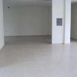 Puste, jasne pomieszczenie z beżowymi płytkami podłogowymi i białymi ścianami, widoczna skrzynka elektryczna na filarze.