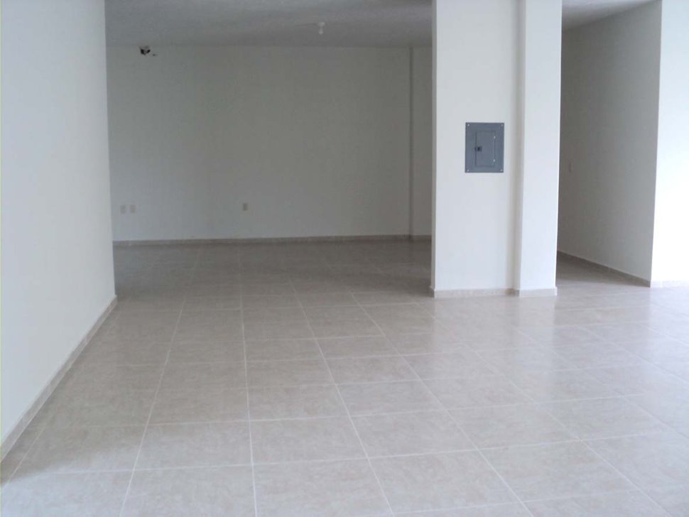 Puste, jasne pomieszczenie z beżowymi płytkami podłogowymi i białymi ścianami, widoczna skrzynka elektryczna na filarze.