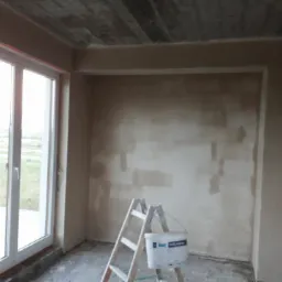 Wnętrze w stanie deweloperskim z nałożonym tynkiem, widoczna drabina oparta o ścianę i wiadro farby, duże okno z widokiem na zewnątrz, surowy betonowy strop.