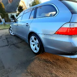 Srebrne kombi BMW na błotnistej drodze wiejskiej, widok z tyłu, z widocznymi śladami opon i odbiciem słońca na karoserii.