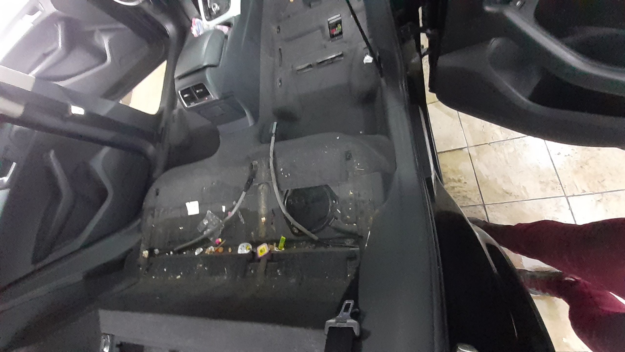 Wnętrze samochodu przed praniem tapicerki, widoczny brud, śmieci i zdemontowane elementy. Otwarta klapa bagażnika i drzwi ukazują fragment podłogi wyłożonej jasnymi płytkami.