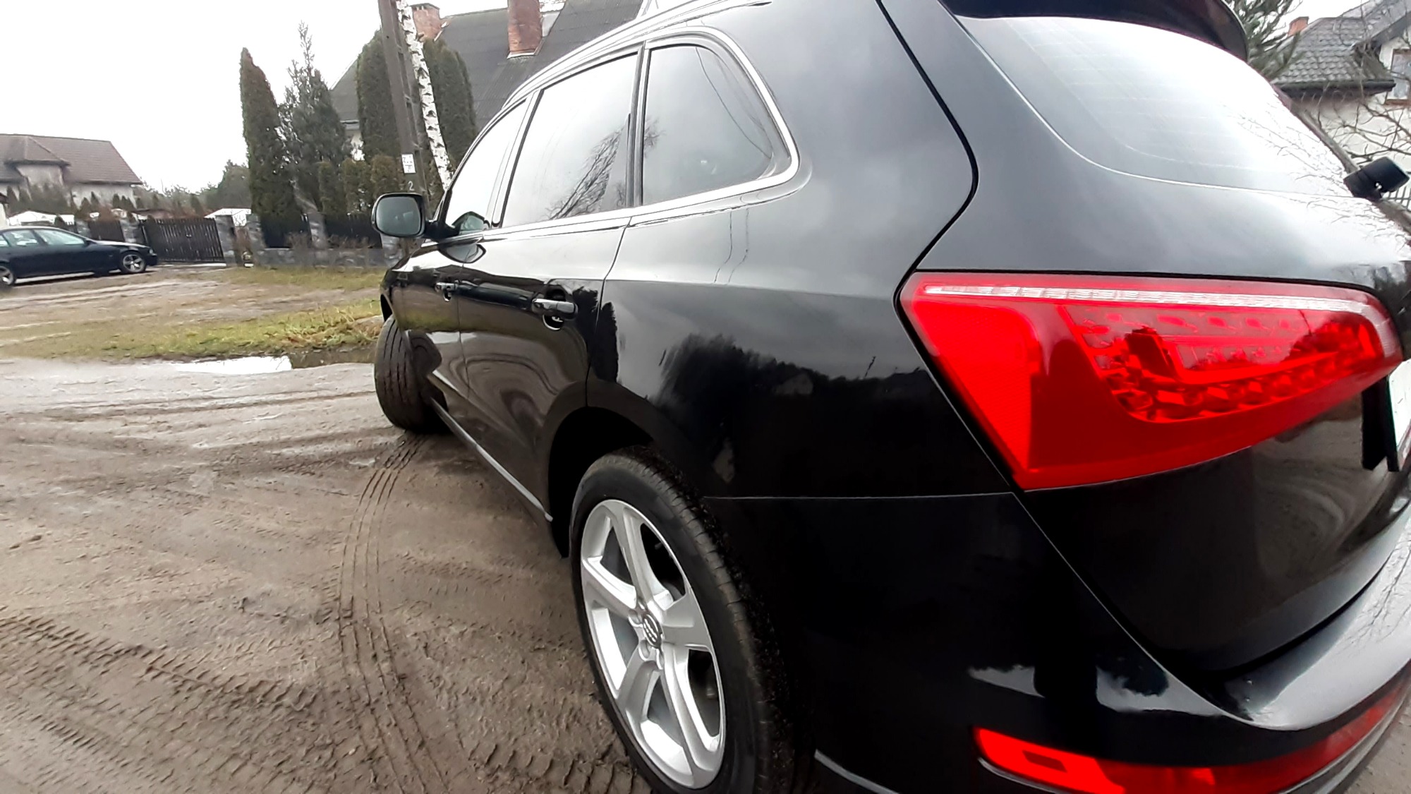 Częściowy widok tyłu czarnego samochodu typu SUV z aluminiowymi felgami na błotnistym podłożu, widoczne odbicia w lakierze.