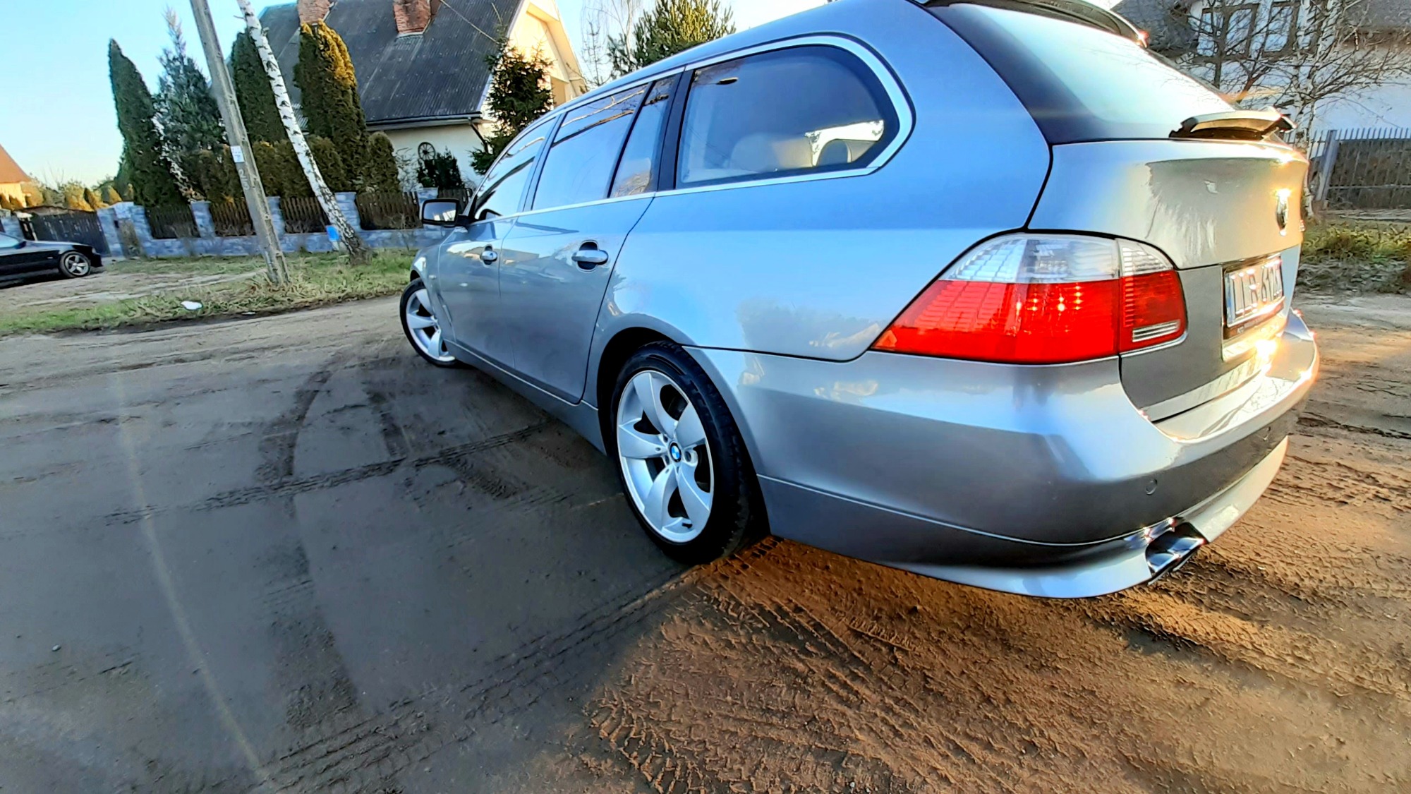 Srebrne kombi BMW na błotnistej drodze wiejskiej, widok z tyłu, z widocznymi śladami opon i odbiciem słońca na karoserii.