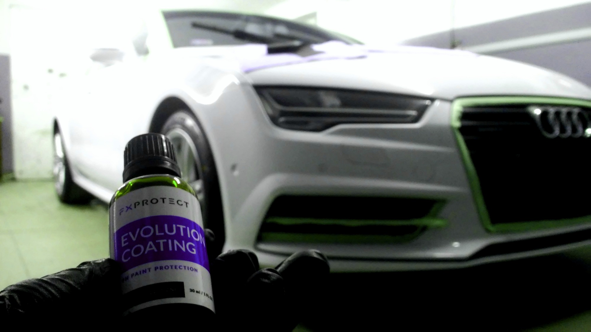 Białe Audi A7 z zieloną taśmą maskującą na grillu, w tle warsztatu, z przodu butelka produktu FX Protect Evolution Coating trzymana w rękawicy.