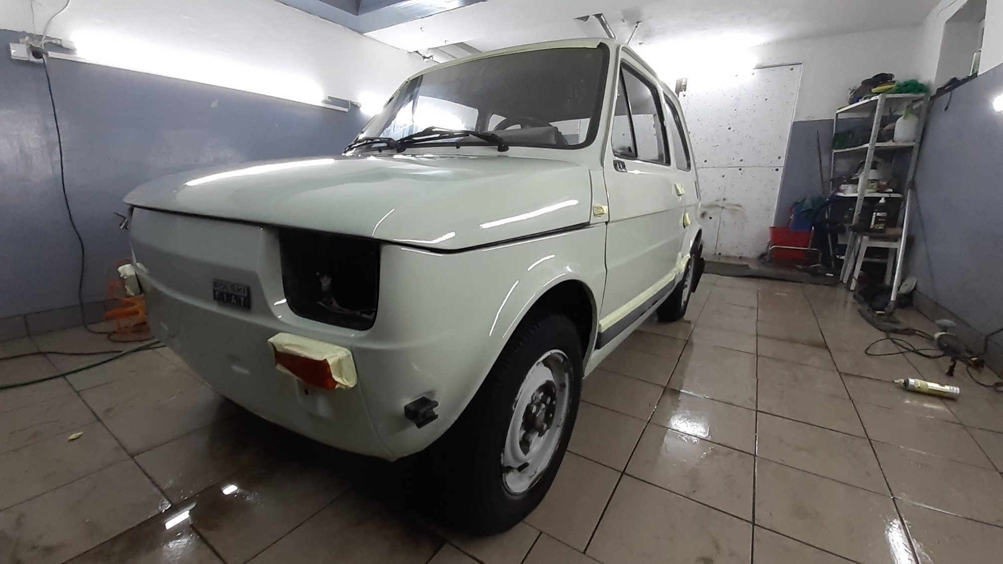 Odrestaurowany, biały Fiat 126p po lakierowaniu, z elementami zabezpieczonymi taśmą malarską, widoczny w warsztacie z błyszczącą podłogą.