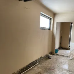 Wnętrze pomieszczenia w trakcie prac wykończeniowych: ściany pokryte świeżym, jasnym tynkiem, widoczne ślady wilgoci przy podłodze, surowy beton na podłodze i aluminiowa listwa pod sufitem.