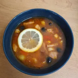 Porcja zupy solianka w ciemnoniebieskiej miseczce, udekorowana plasterkiem cytryny i czarnymi oliwkami, na drewnianym stole.