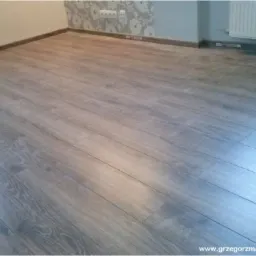 Wykończone wnętrze z panelami podłogowymi w odcieniach szarości, listwy przypodłogowe w kolorze drewna, ściana z tapetą w kwiaty i grzejnik.