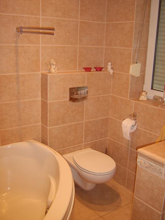 Jasna łazienka z beżowymi płytkami, widoczna wanna narożna, podwieszana toaleta, półka z dekoracjami aniołków i czerwonymi miseczkami, oraz grzejnik drabinkowy na ręczniki.