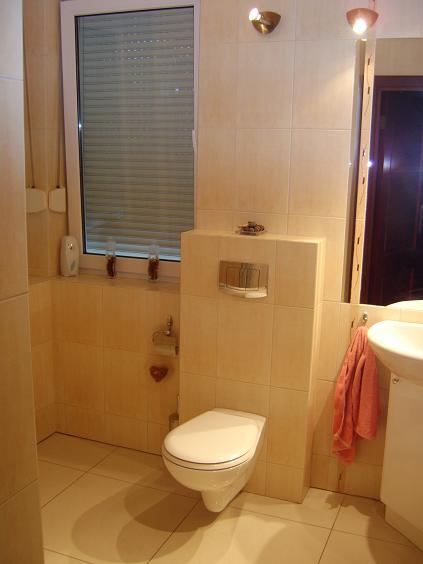 Jasna łazienka z beżowymi płytkami, wisząca toaleta, okno z roletą i akcent w postaci czerwonego serduszka na ścianie.