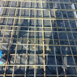 Siatka zbrojeniowa przygotowana na czarnej folii, z widocznymi elementami dystansowymi i fragmentem niebieskiej folii ochronnej w rogu.