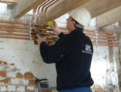 Instalator w kasku lutuje miedziane rury centralnego ogrzewania w surowym wnętrzu, widoczne drewniane belki stropowe i fragment ceglanej ściany.