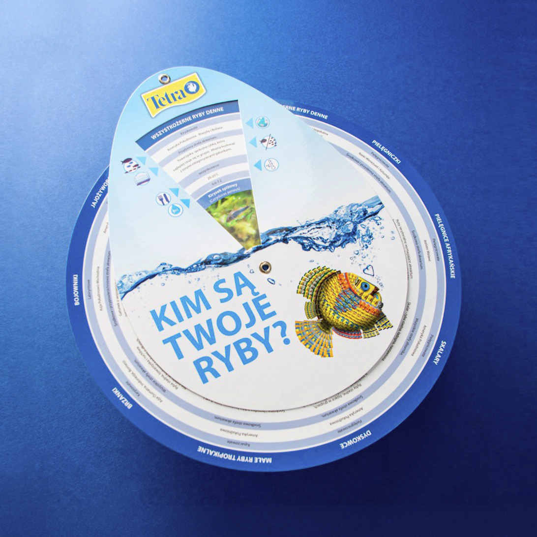 Okrągła, interaktywna zawieszka informacyjna marki Tetra z pytaniem 'Kim są Twoje ryby?', prezentująca różne gatunki ryb akwariowych i ich charakterystykę na niebieskim tle.