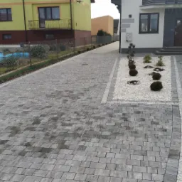 Szeroki podjazd z szarej kostki brukowej prowadzący do domu, z obrzeżami z jaśniejszej kostki, rabaty z białym żwirem i kulistymi krzewami po bokach.