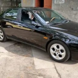 Czarny samochód osobowy marki Alfa Romeo zaparkowany na betonowym podjeździe, widoczny z boku, z charakterystycznymi felgami i drewnianą kierownicą.