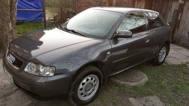 Szary samochód Audi A3 zaparkowany na trawie obok betonowego chodnika i studzienki kanalizacyjnej, widoczne zabrudzenia na karoserii i oponach.