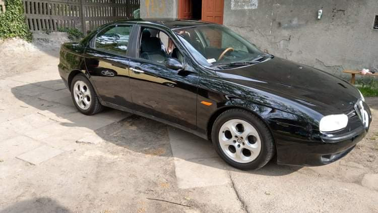 Czarny samochód osobowy marki Alfa Romeo zaparkowany na betonowym podjeździe, widoczny z boku, z charakterystycznymi felgami i drewnianą kierownicą.