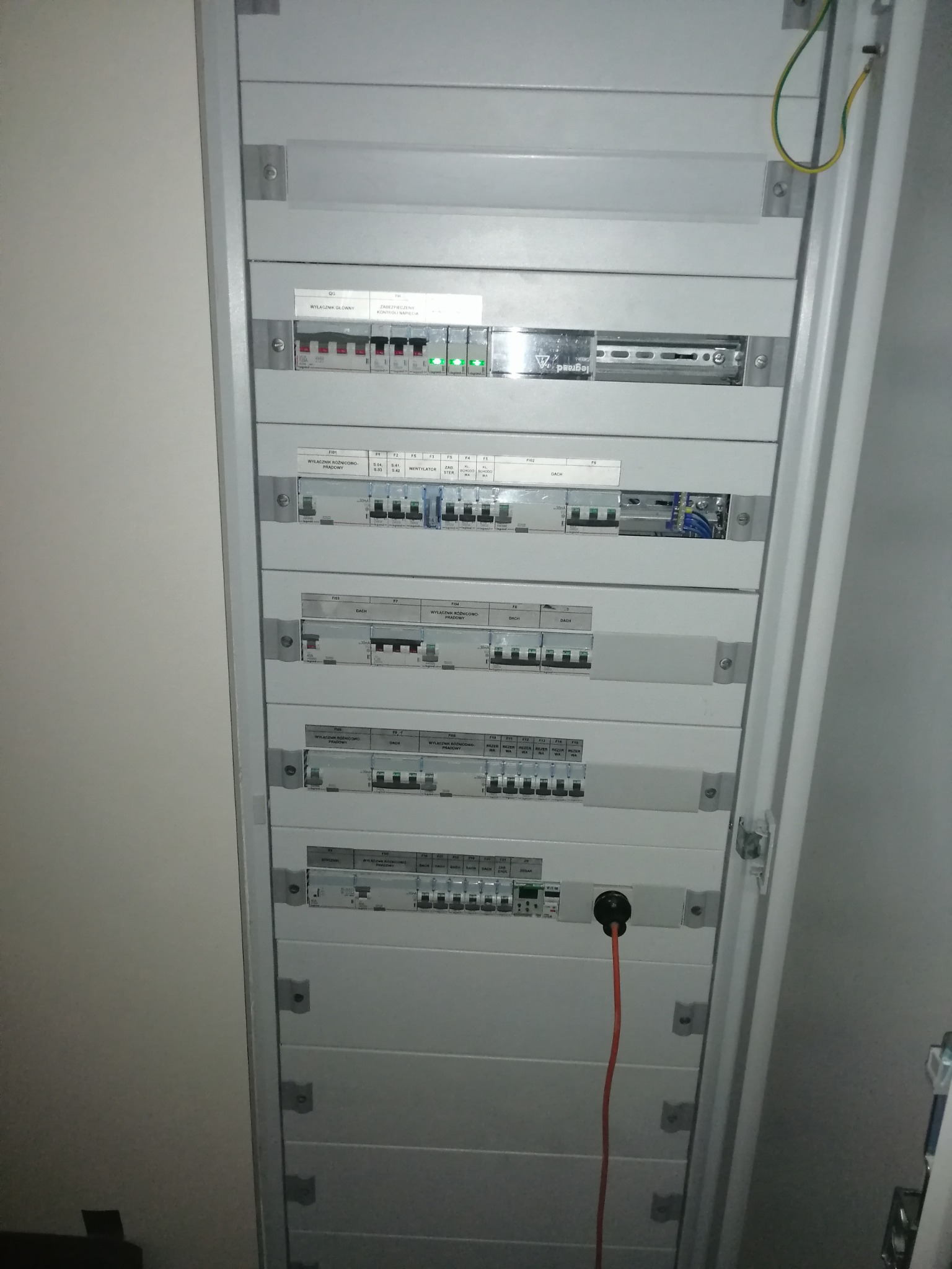 Otwarta szara skrzynka rozdzielcza z zainstalowanymi bezpiecznikami i wyłącznikami, widoczny kabel zasilający z czarną wtyczką podłączony do jednego z obwodów.