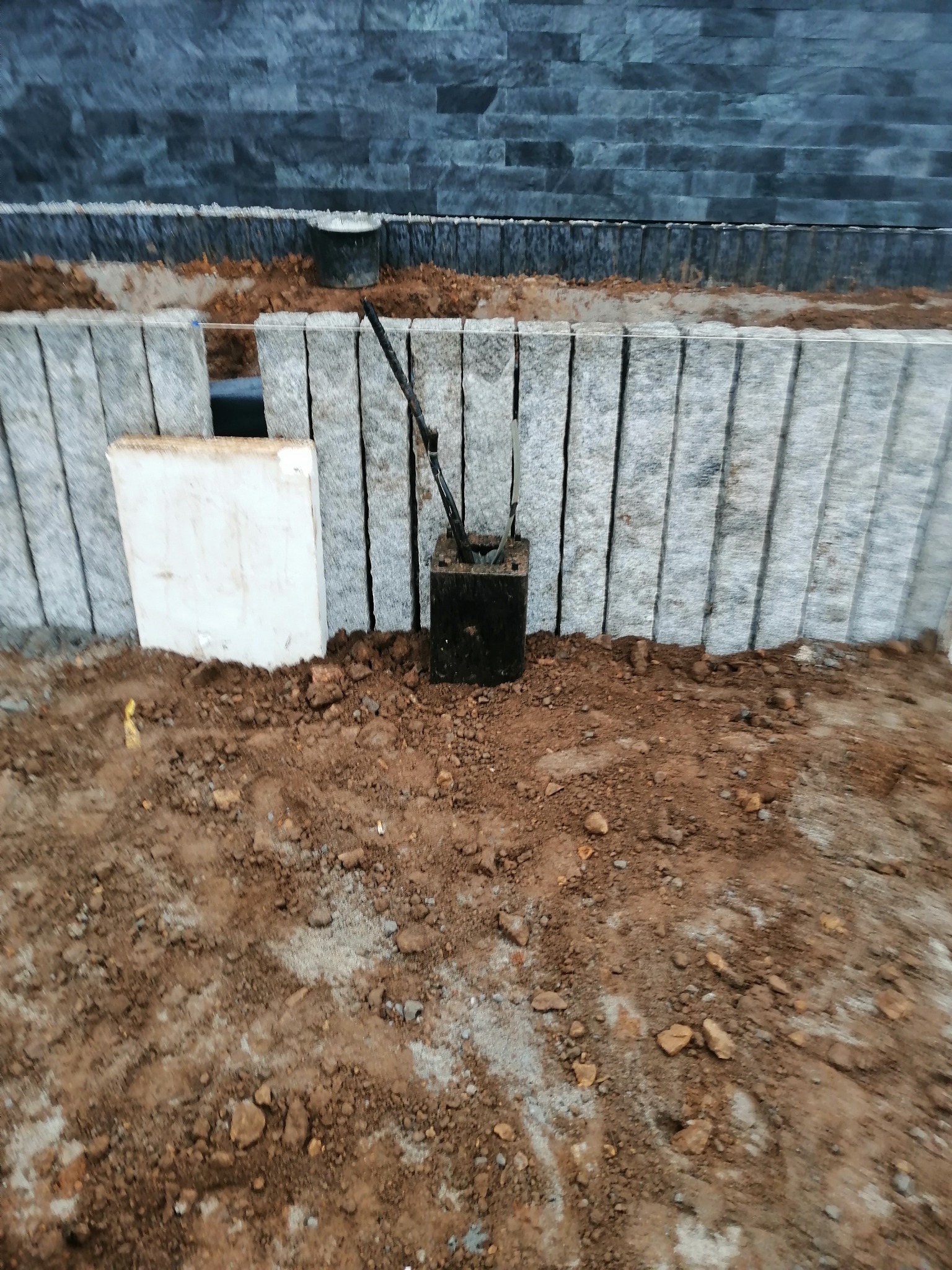 Czarna puszka instalacyjna z wystającymi przewodami, umieszczona w ziemi przy nowo wzniesionym, szarym murku z kamiennych bloków, na tle ciemnej ściany z kamienia łupanego.