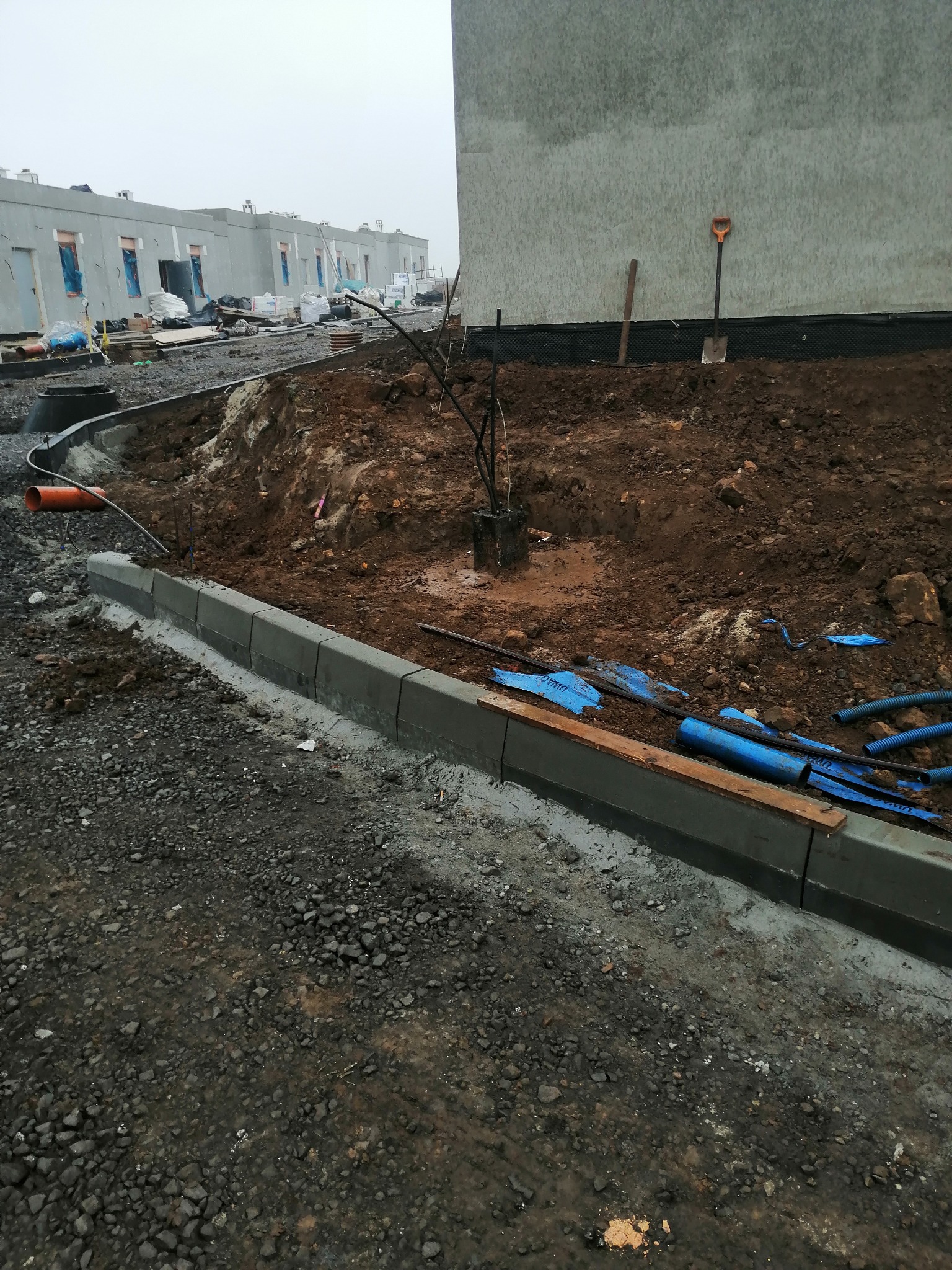 Nowe budynki w tle, widoczne kable i rury w wykopie, świeżo ułożony betonowy krawężnik oddzielający chodnik od ziemi.
