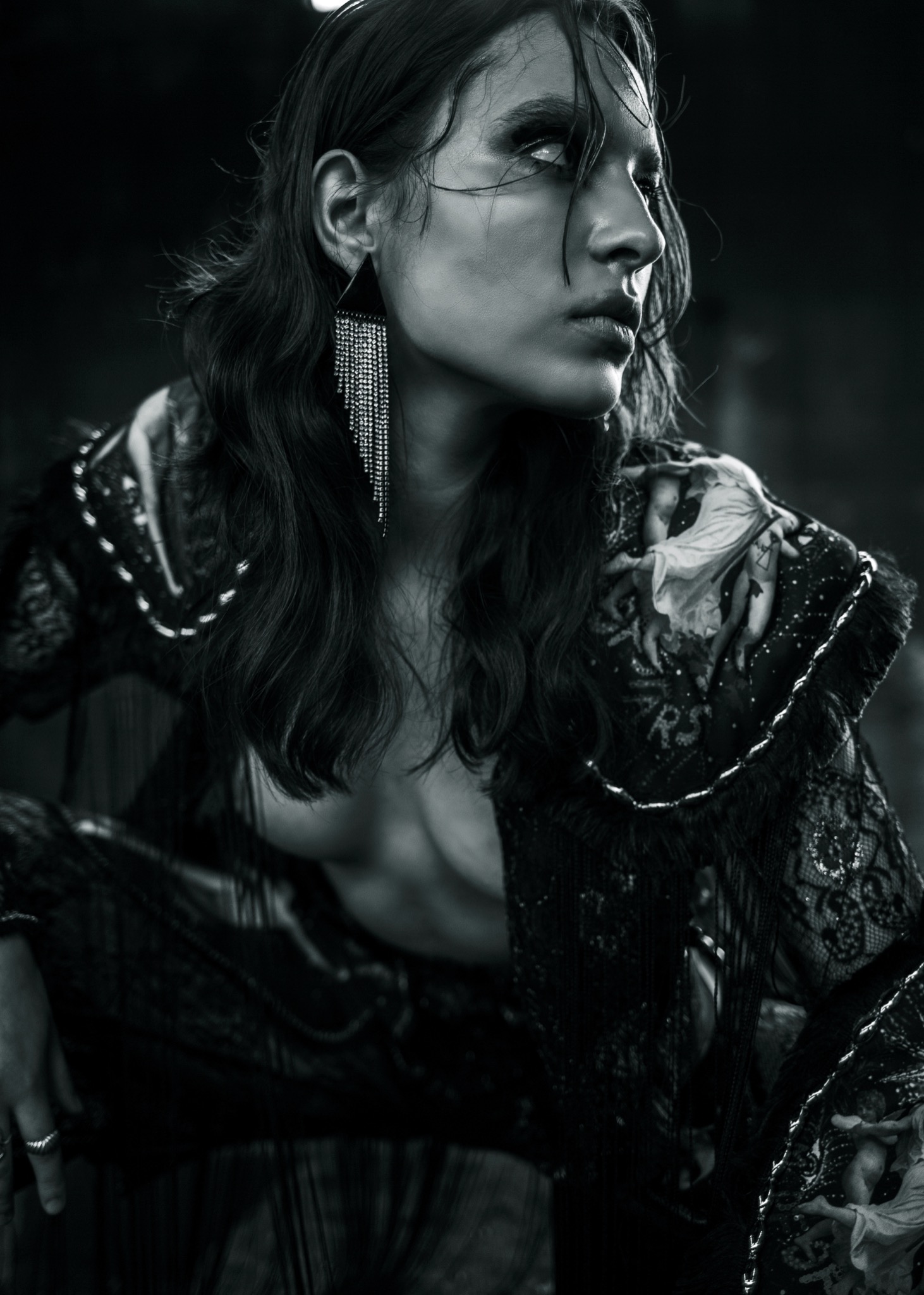 Czarno-białe, artystyczne portretowe ujęcie kobiety z mokrymi włosami, w ozdobnym stroju z frędzlami i dużym, błyszczącym kolczykiem, spoglądającej w górę.