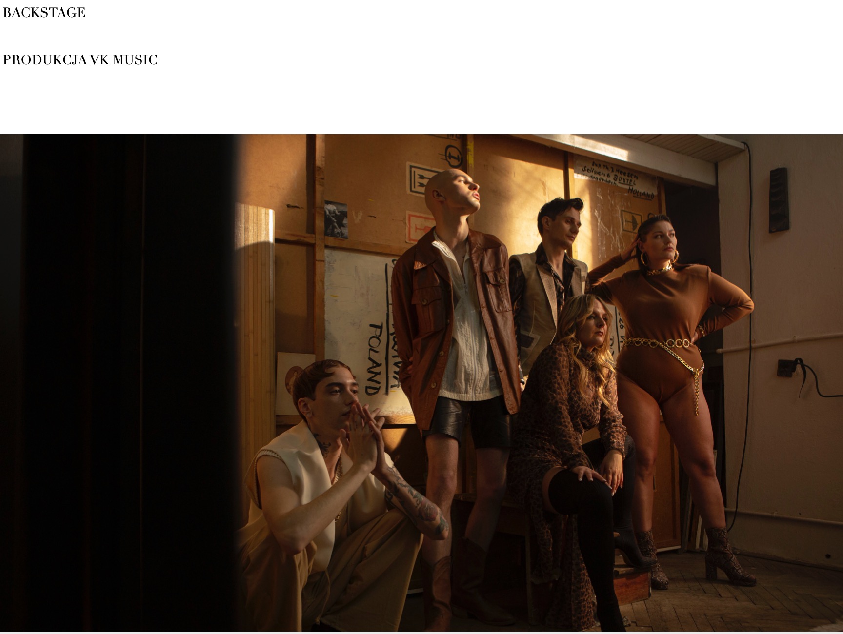 Grupa pięciu osób w różnorodnych stylizacjach pozuje w pomieszczeniu z surowymi ścianami. Mężczyzna w skórzanej kurtce i krótkich spodenkach stoi obok kobiety w brązowym body z łańcuchem. Z przodu...