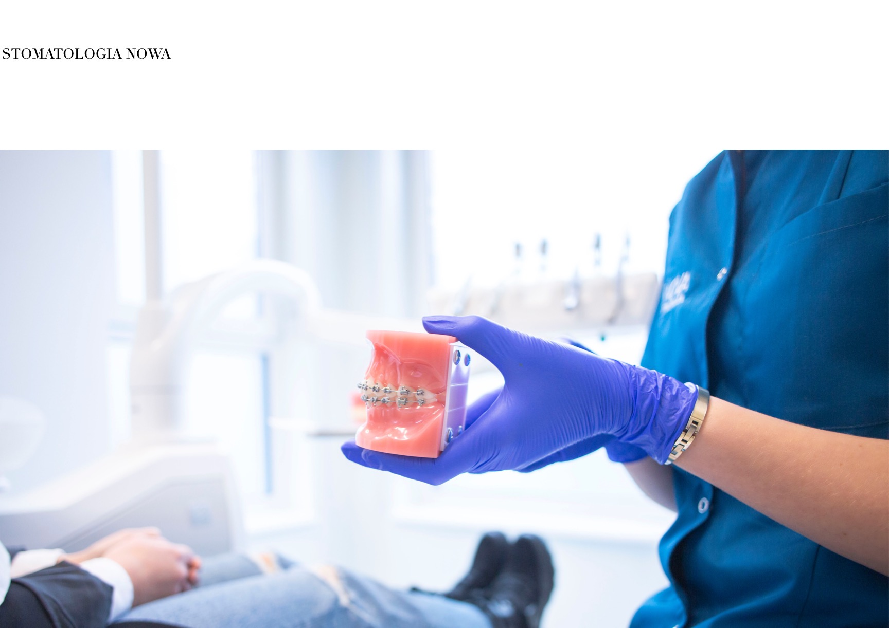 Dentysta w niebieskich rękawiczkach trzyma model szczęki z założonym aparatem ortodontycznym, w tle widoczny fotel dentystyczny i pacjent.