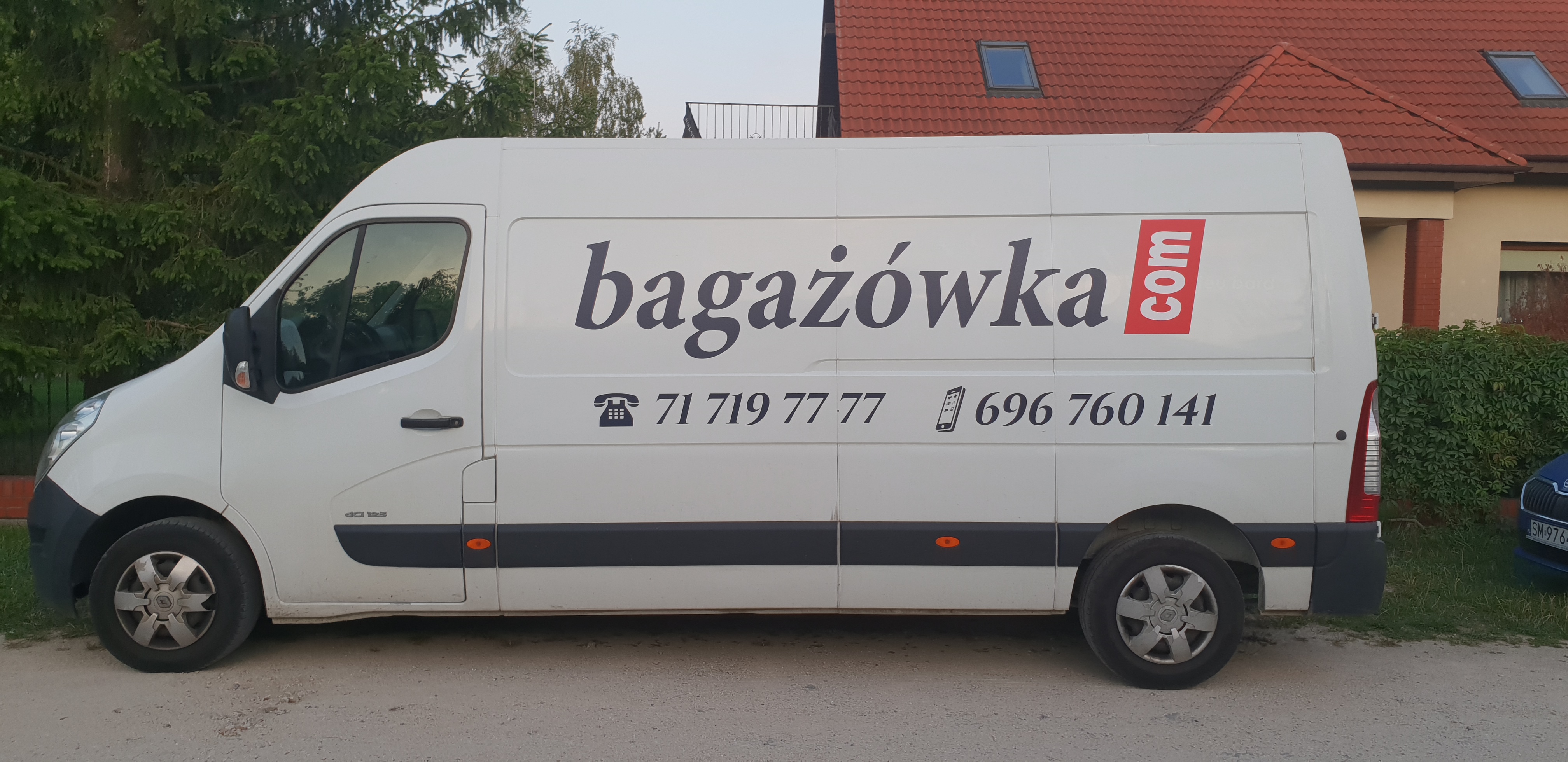 Biały samochód dostawczy z logo firmy bagażówka.com i numerami telefonów zaparkowany na żwirowej drodze przed domem.