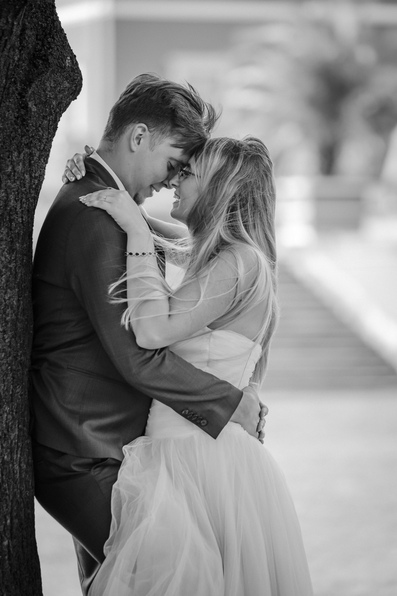 Czarno-białe zdjęcie obejmującej się pary młodej, opierającej się o drzewo, z delikatnym uśmiechem na twarzy panny młodej i widocznym pierścionkiem zaręczynowym.