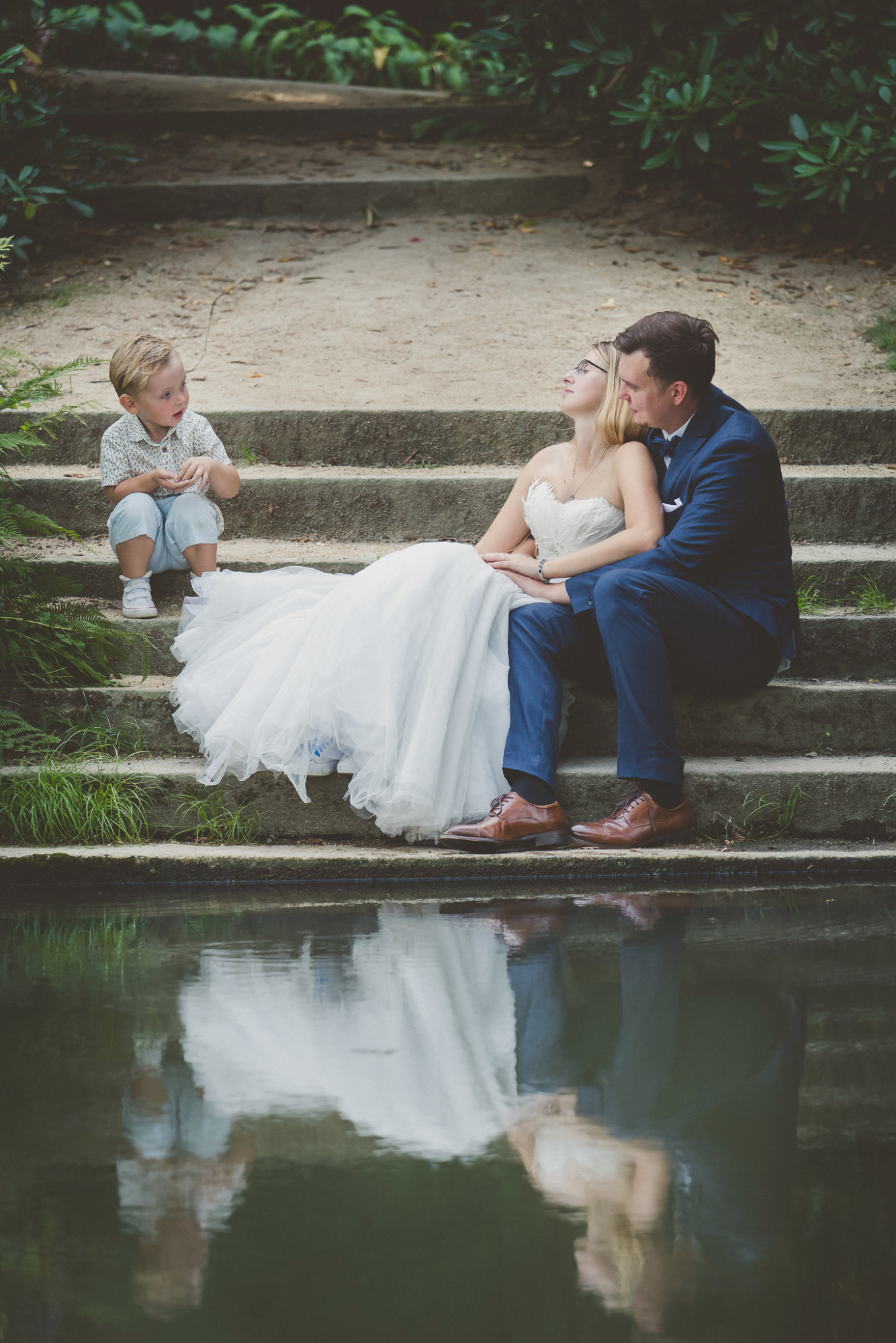 Para młoda w strojach ślubnych siedzi na kamiennych schodach obok stawu, z odbiciem w wodzie i małym chłopcem siedzącym obok, zieleń w tle.