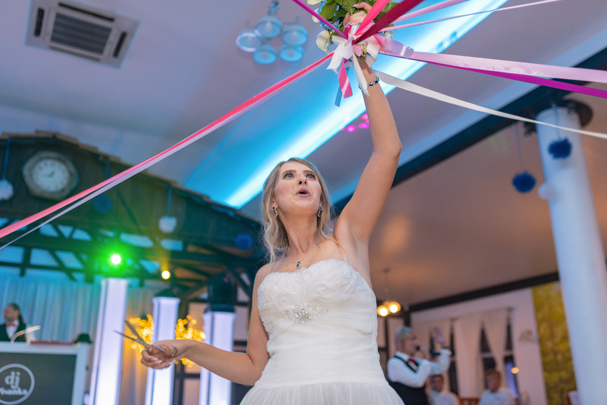 Panna młoda w białej sukni ślubnej, unosząca bukiet z kolorowymi wstążkami podczas oczepin, z nożyczkami w dłoni, w tle DJ i goście weselni.